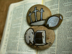 Steine, die auf einer Bibelseite liegen und bemalt sind mit drei Kreuzen, dem geöffneten Grab Jesu und einem Fisch (Ichthys)